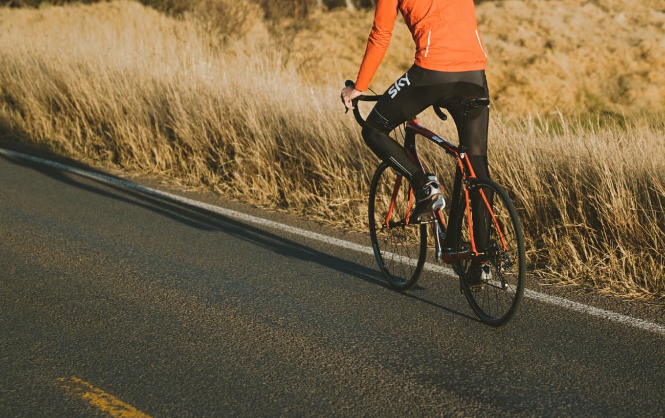 A cyclist in an orange jersey rides on a tarmac road past a wheat field.