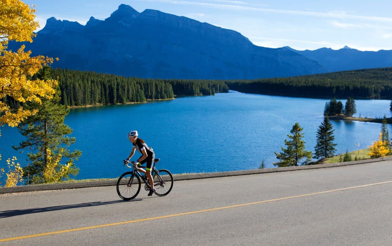 A cyclist rides past a cool blue lake with a mountain looming behind.