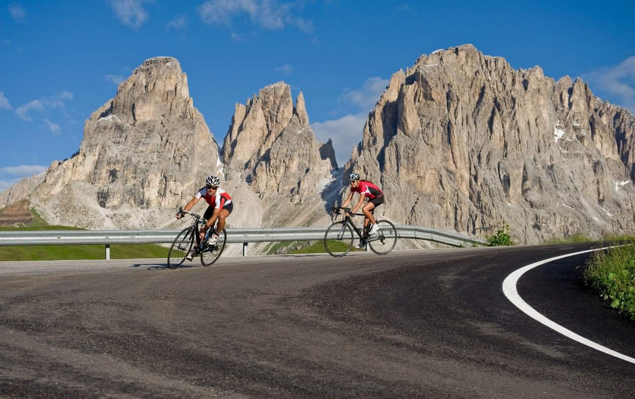 Two cyclists descend a hairpin bend with a jagged mountain range behind them.