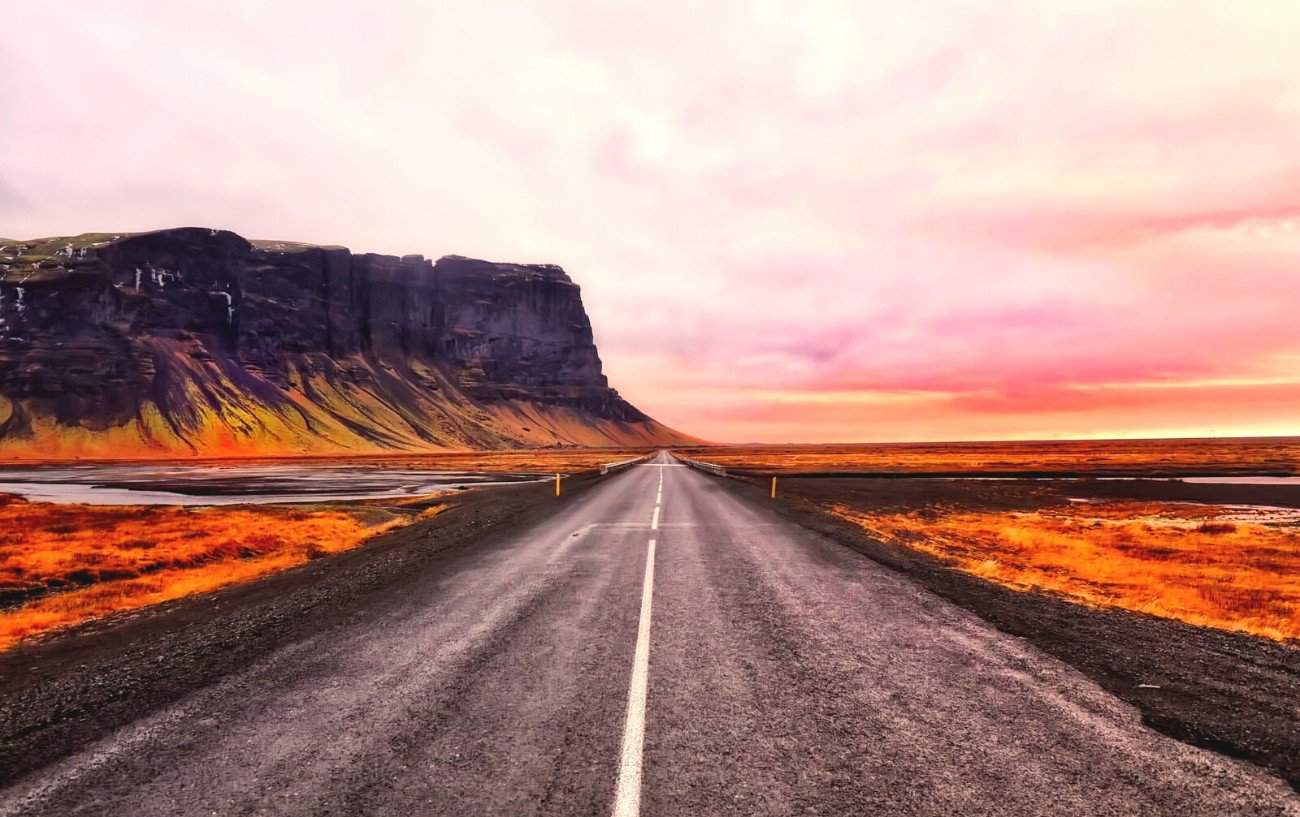 Icelandic road at sunset, lit up with blazing orange and pink colours against volcanic black cliffs.