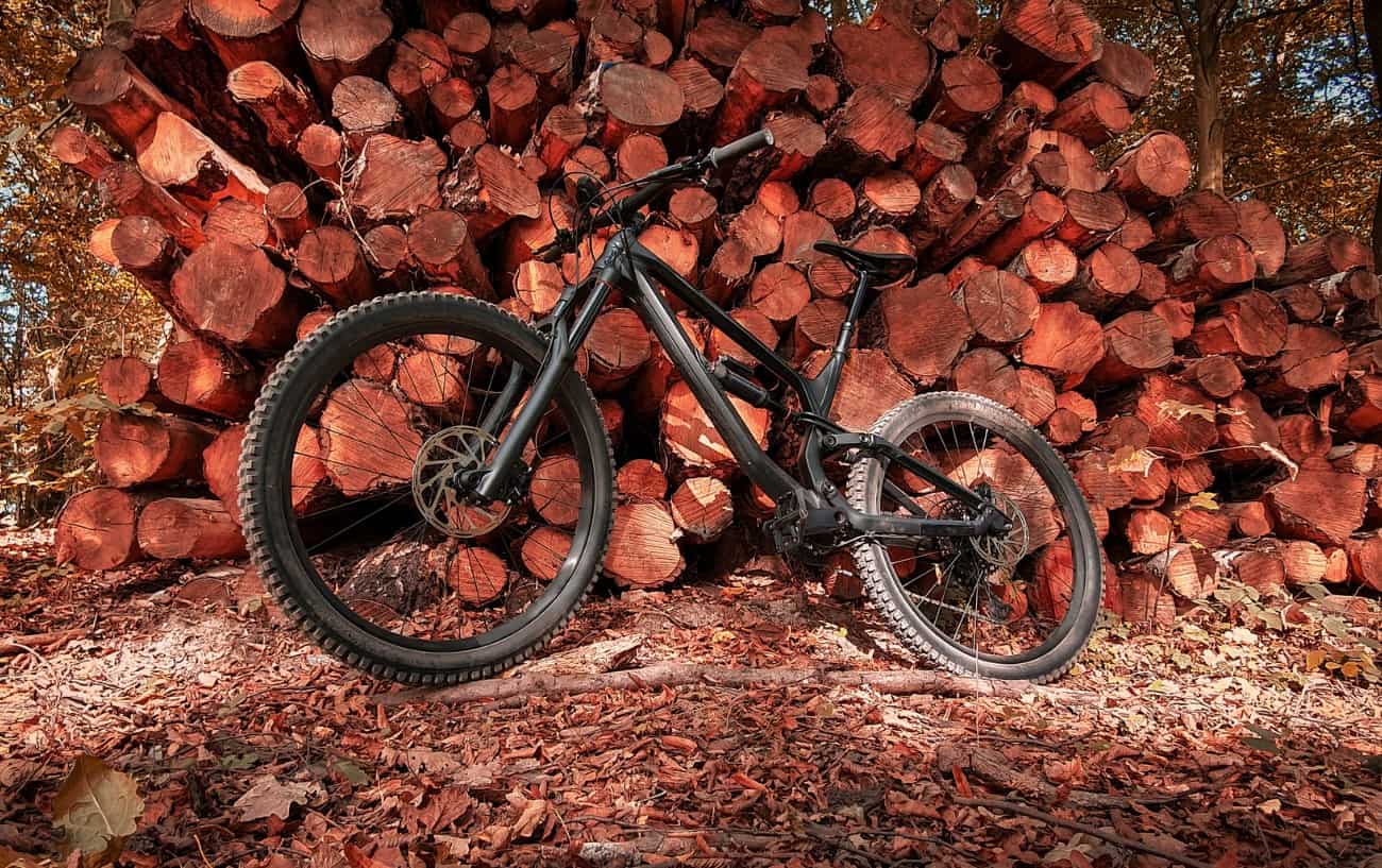 A black full suspension bike leans against a log pile in an autumnal setting.