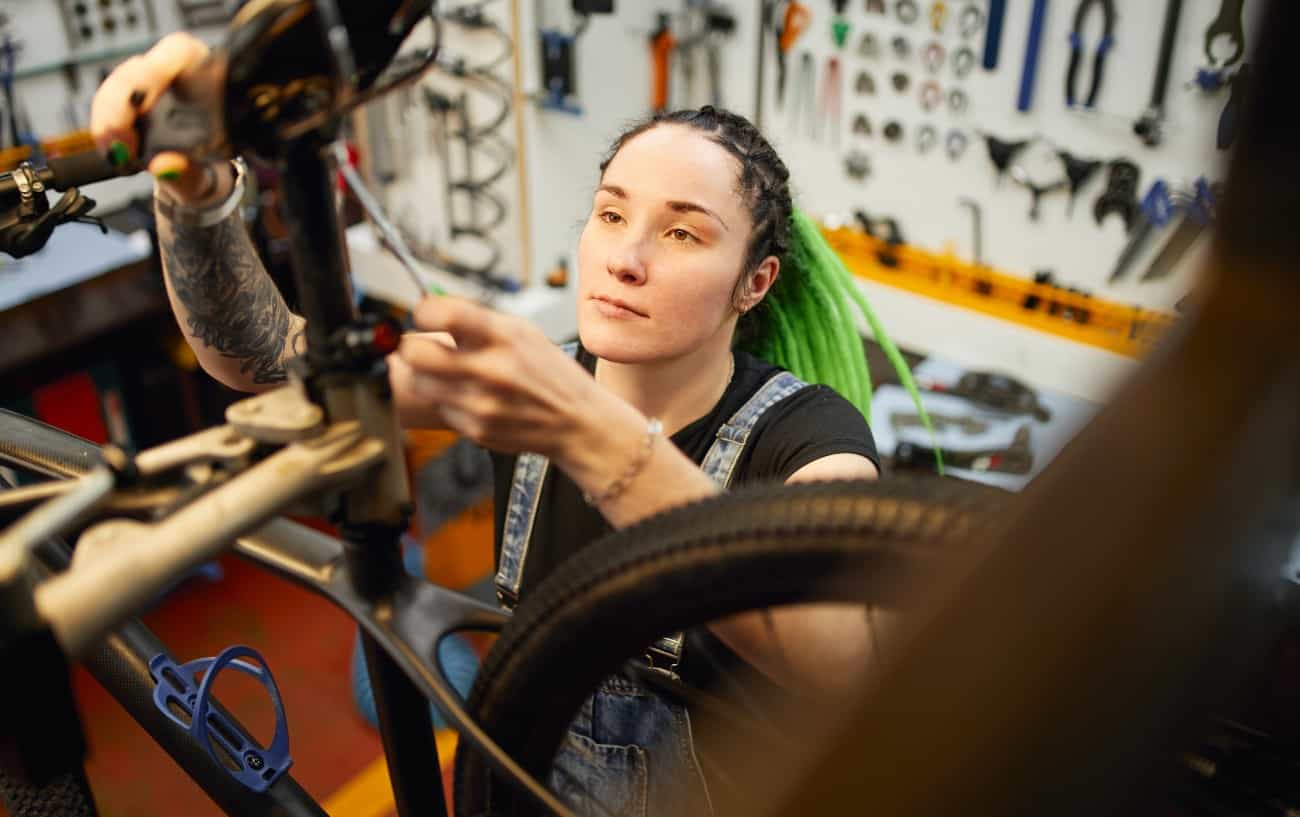 A mechanic with green hair adjusts her bike's saddle in a workshop.