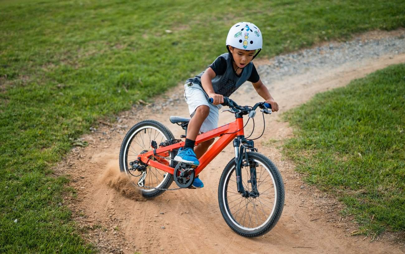 A kid drifts his orange mountain bike around a dirt track.