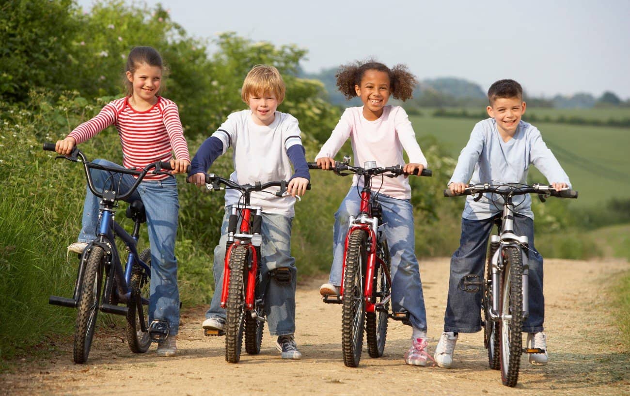 Four kids ride their bikes on a dirt path through a field.