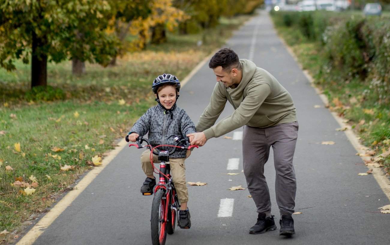 A dad teaches his son to ride a bike on a tarmac path through a park.