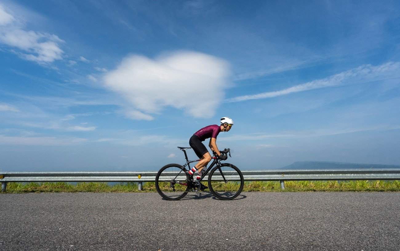 A cyclist in a burgundy jersey pedals along a flat tarmac ride while standing up on a bike.