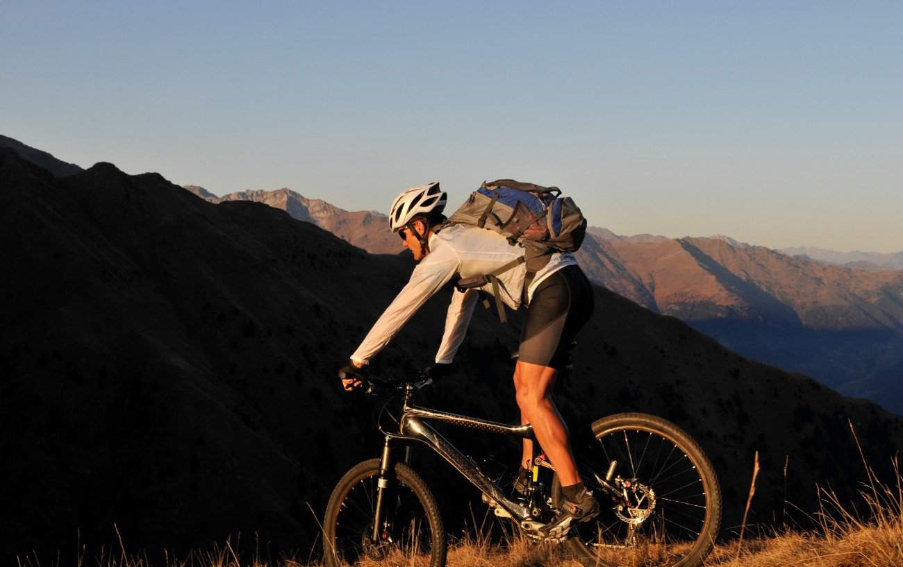 A mountain biker in a grey jersey descends a hill into a sunlit valley.