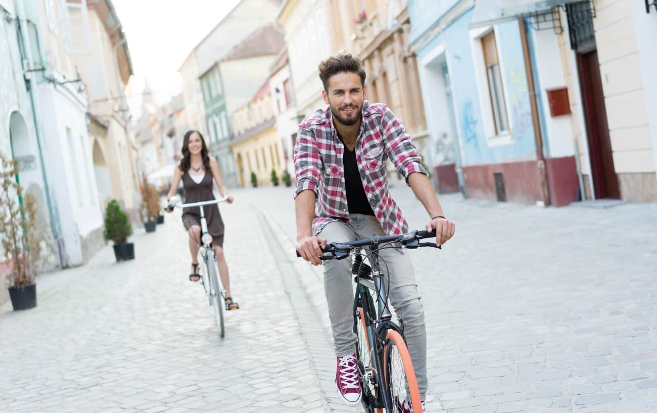 A couple on hybrid bikes cycle down a cobbled street in a European town.