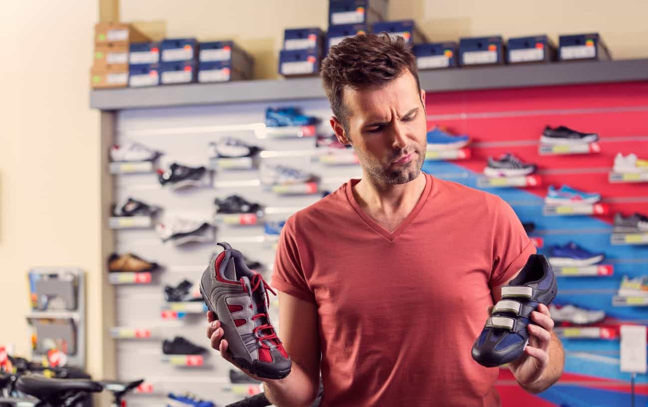 A man in. ared t-shirt compares two different cycling cleats types in a shop.