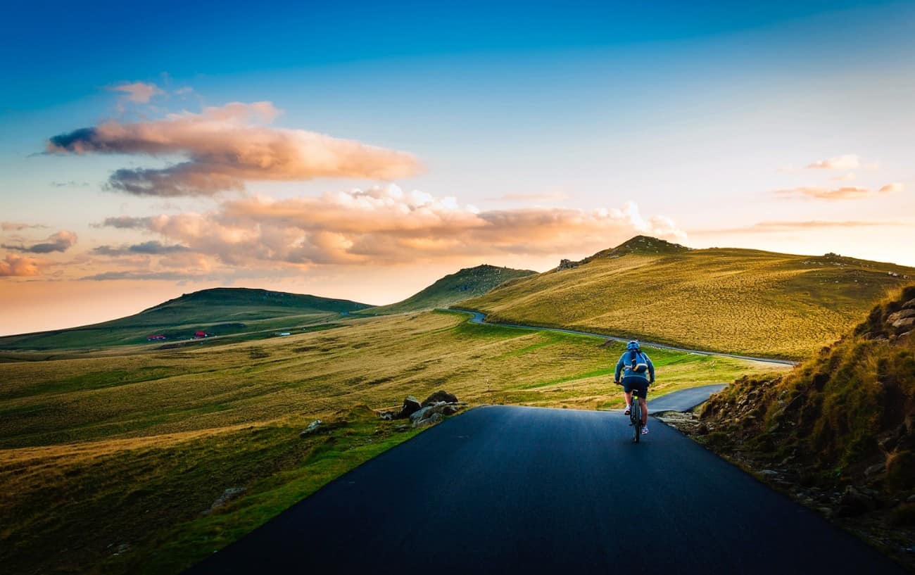 A cyclist in a blue jumper rides along a tarmac road through grassy hills at sunset.