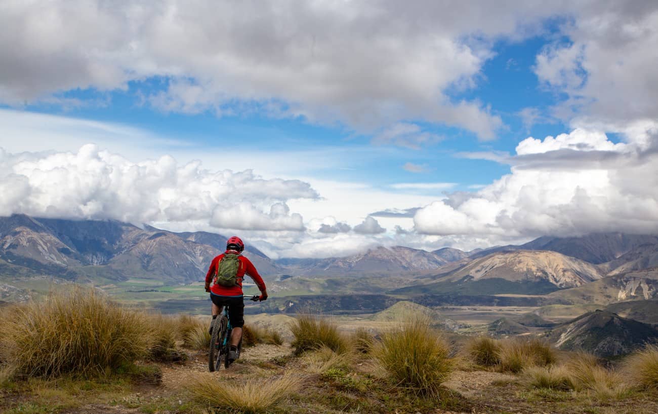 A cyclist in a red jumper rides through grassland toward a mountain vista.