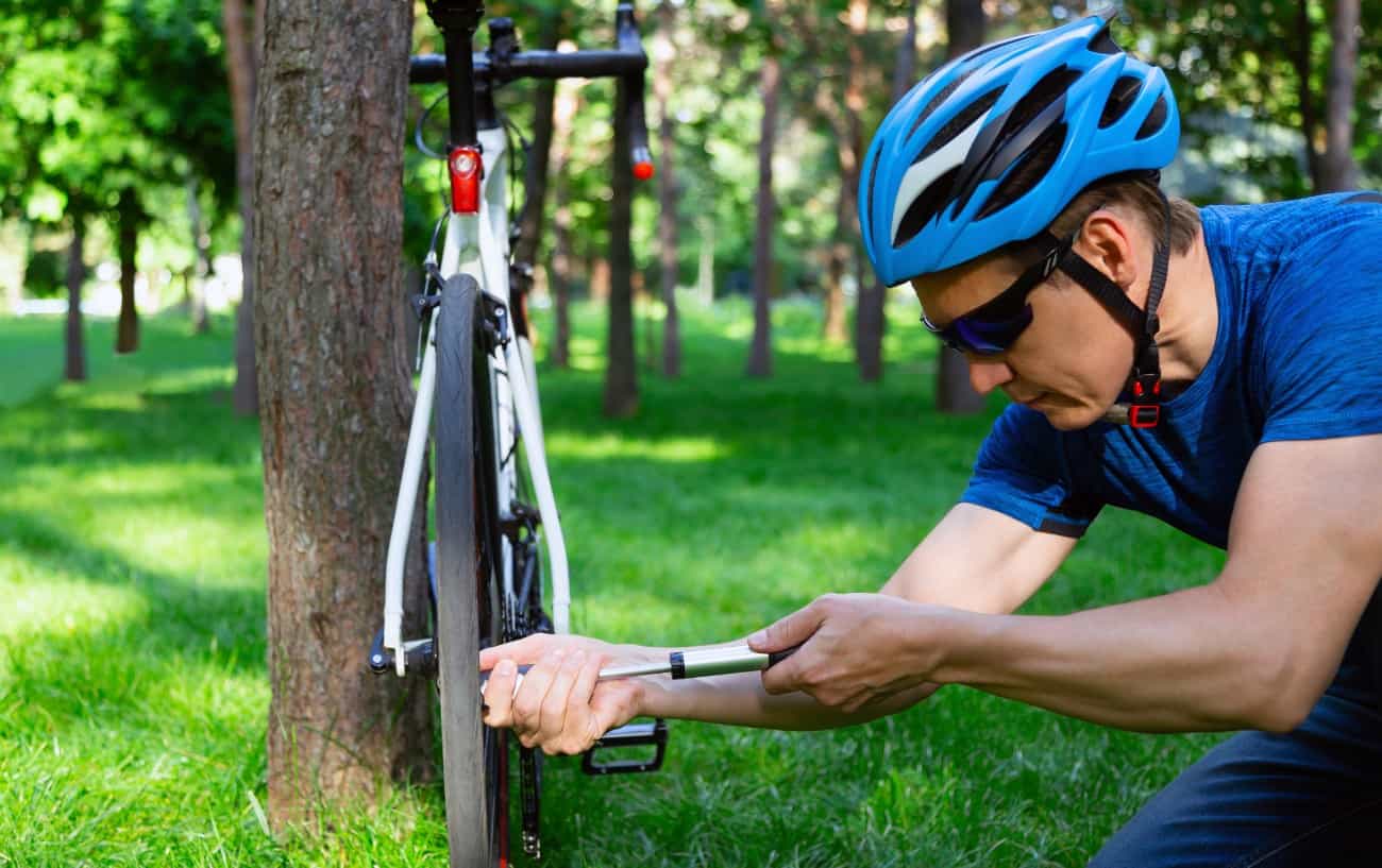 A man in a blue t-shirt and a blue helmet demonstrates how to pump a bike tire in a forest.