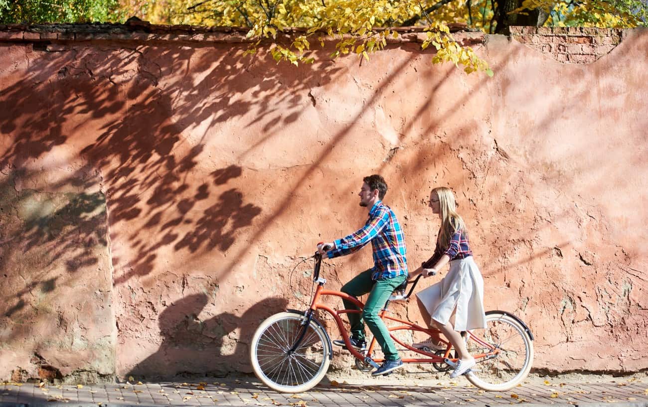 A couple ride an orange tandem bike in front of a terra cotta wall.