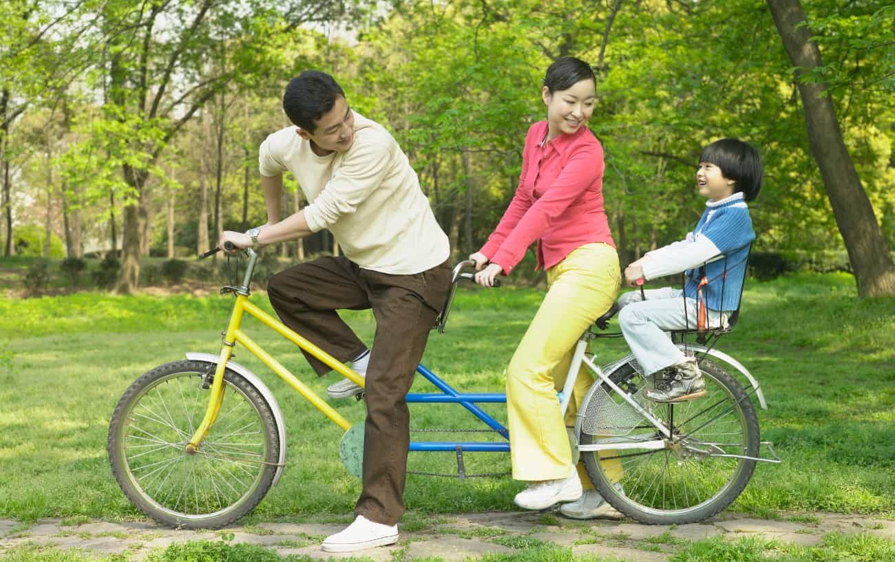 A young family ride a blue and yellow tandem bike through a grassy park.