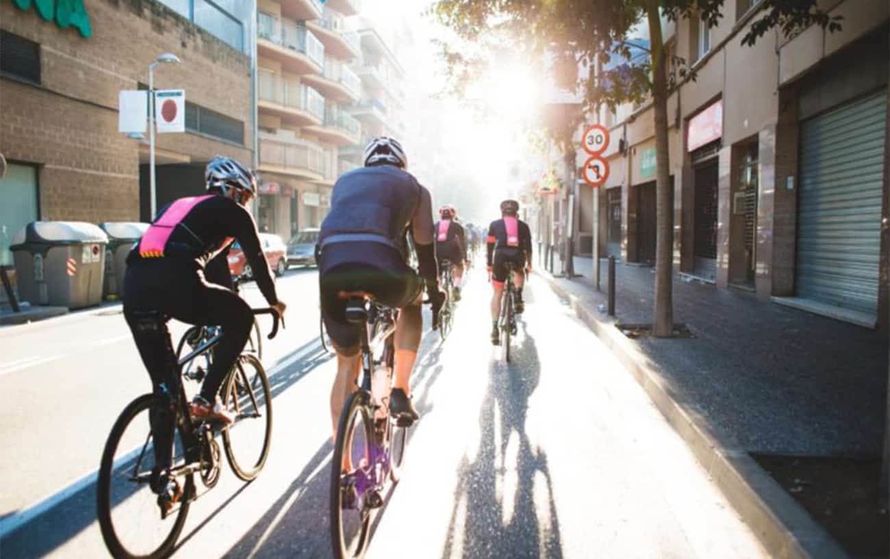 A group of cyclists in black and pink jerseys ride through a city in the afternoon sun.