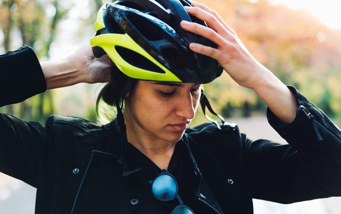 A woman in a black jacket puts on a black bike helmet.