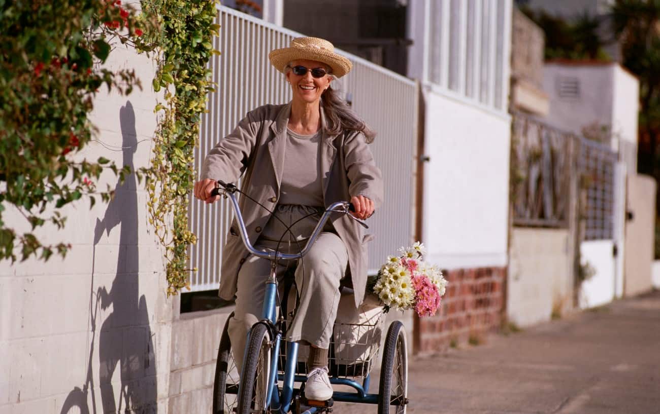 A woman rides her adult trike down the pavement, with flowers in her basket.