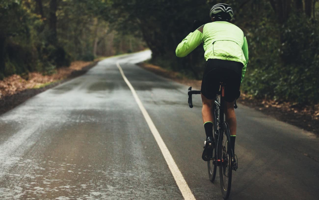 A man in a green raincoat rides a black road bike on a forested road.