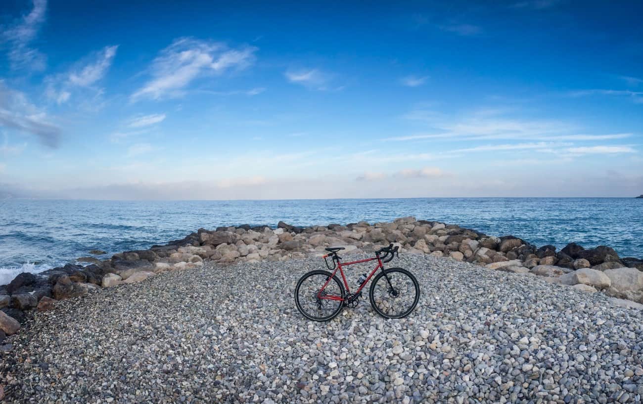 A red gravel bike stands on a pebbled coastline at sunset.