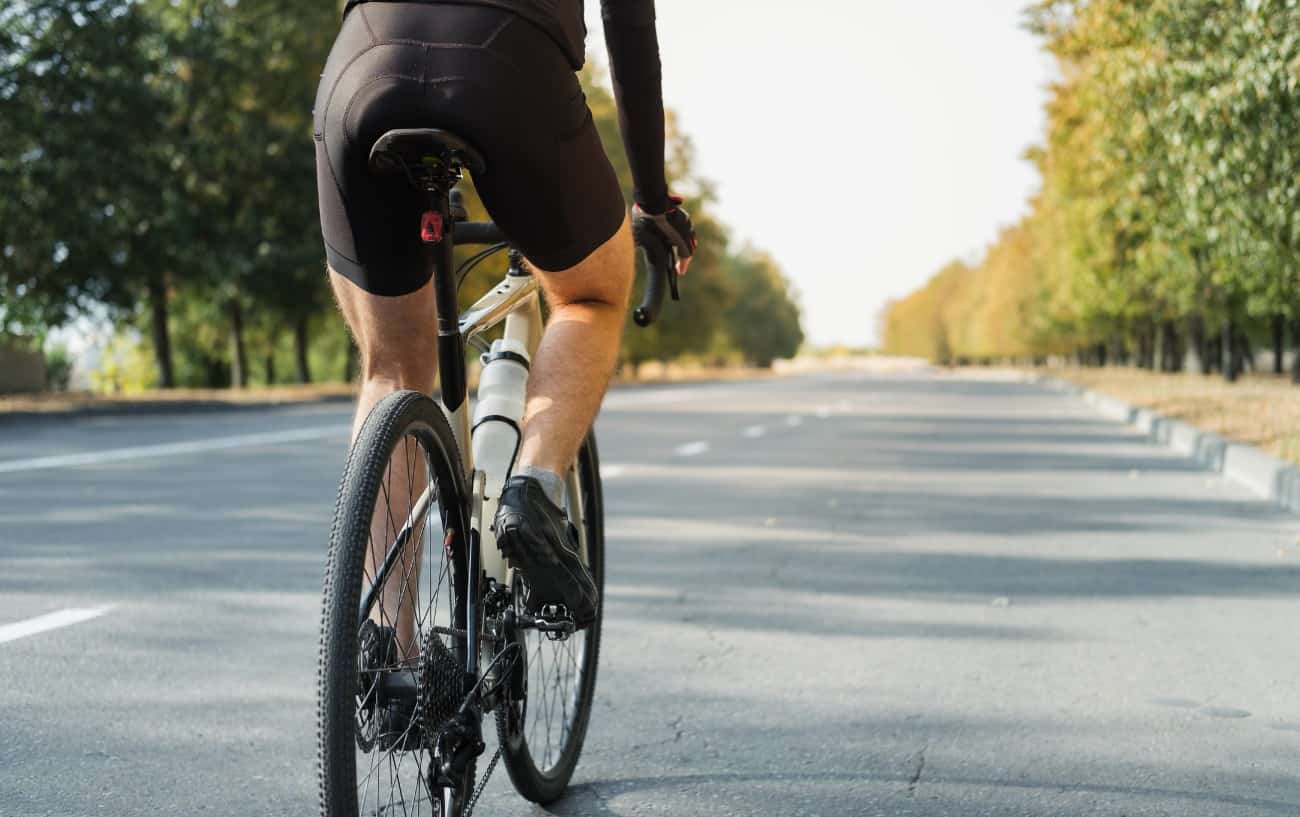 A rider in black cycling shorts rides a white gravel bike on a tarmac road.