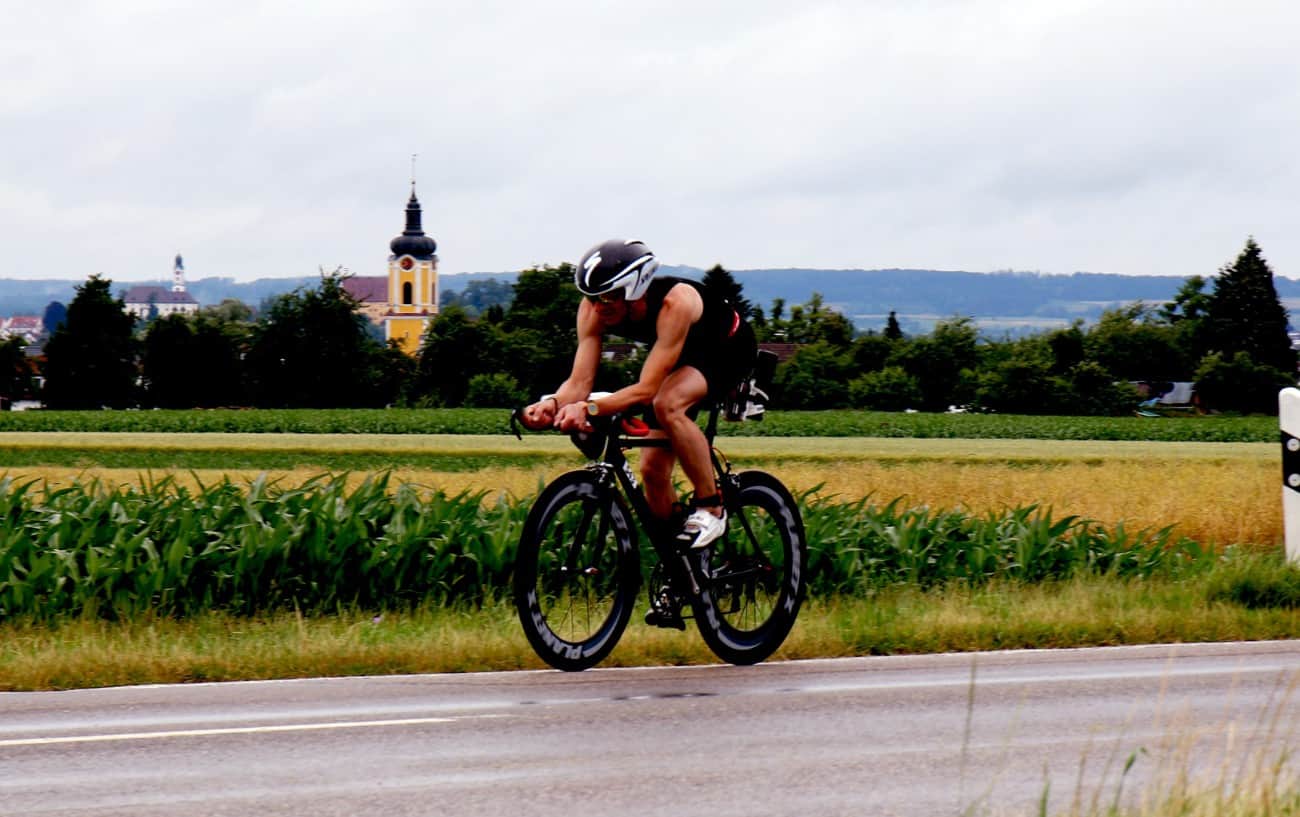 A triathlete in a black jersey rides a black tri bike past a corn field.