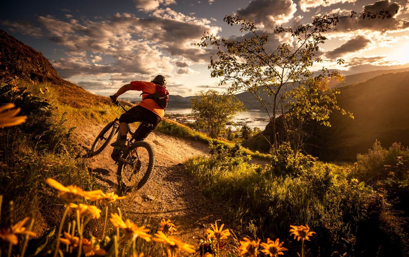 A cyclist on a black cross country bike rides around a steeply banked dirt trail at sunset.