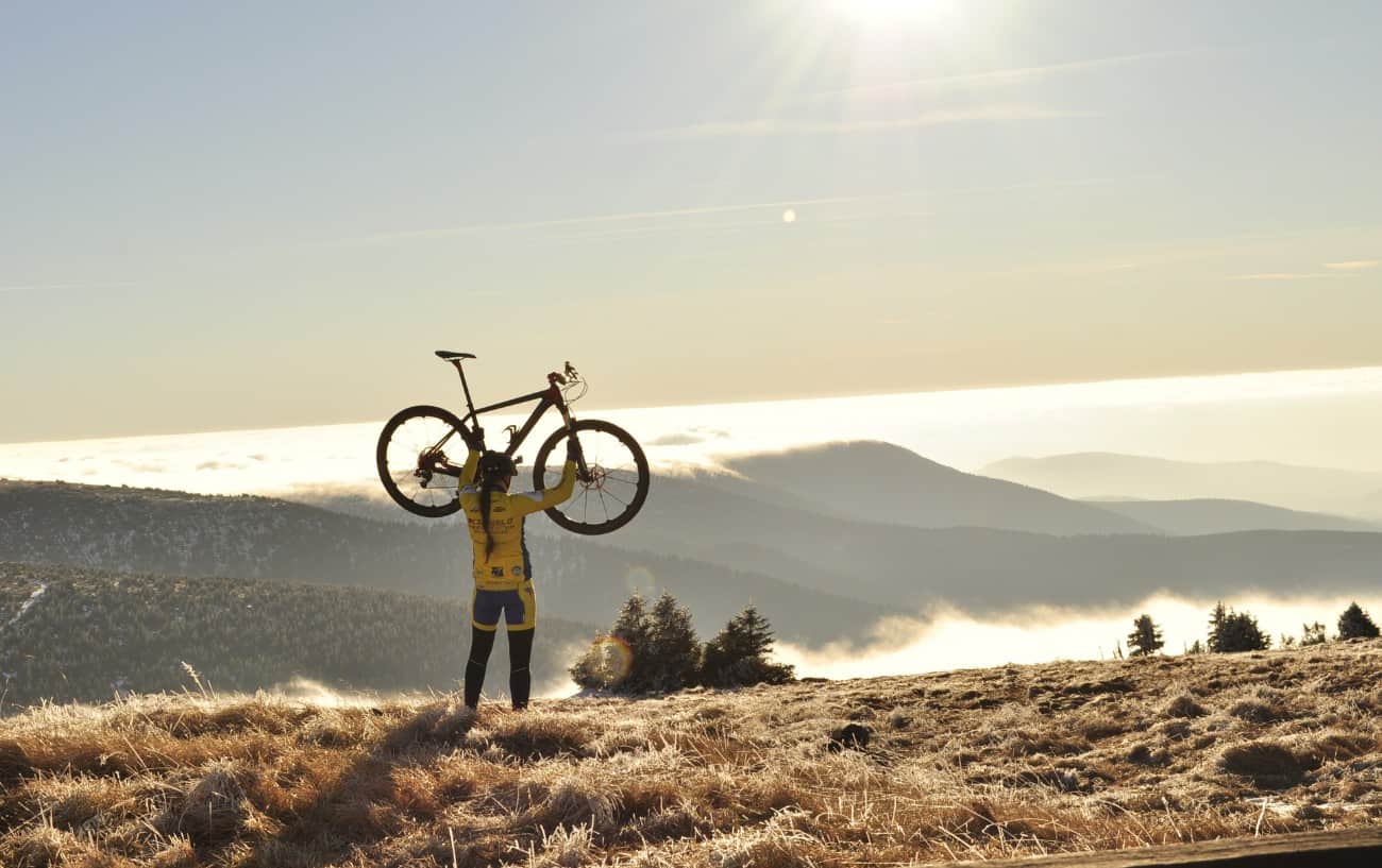A cyclist lifts her mountain bike above her head while looking down into a sun-filled valley.