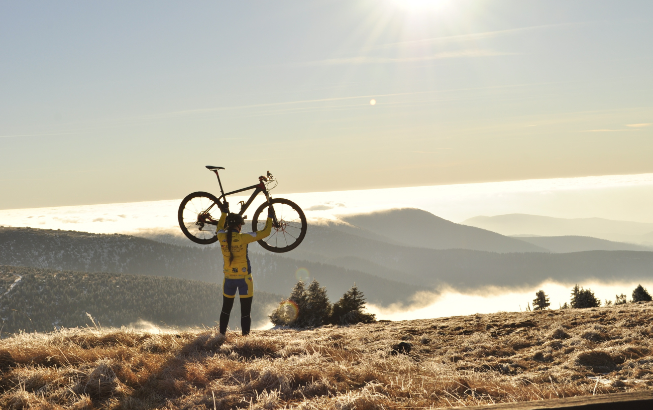 A rider in a yellow jersey holds up her mountain bike in front of a cloud-filled valley.