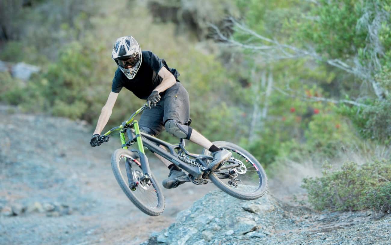 A cyclist on a black downhill mountain bike leaps off a gravel jump.