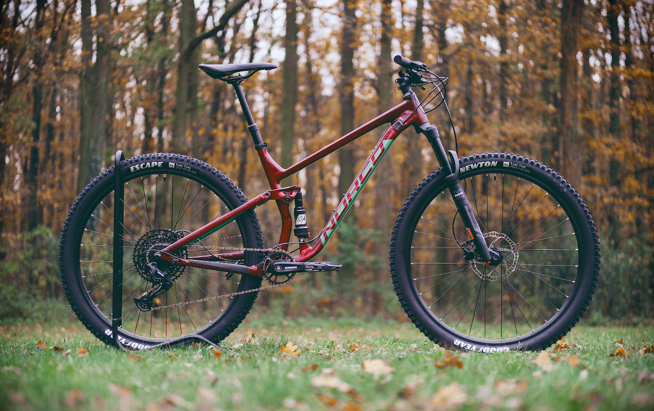 A burgundy enduro mountain bike sits in front of a forest.