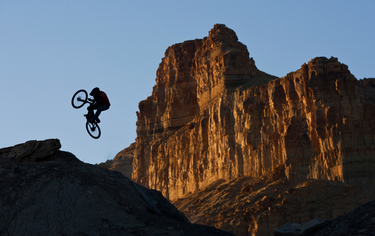Silhouette of a dirt jump bike taking off against a sunlit canyon backdrop.