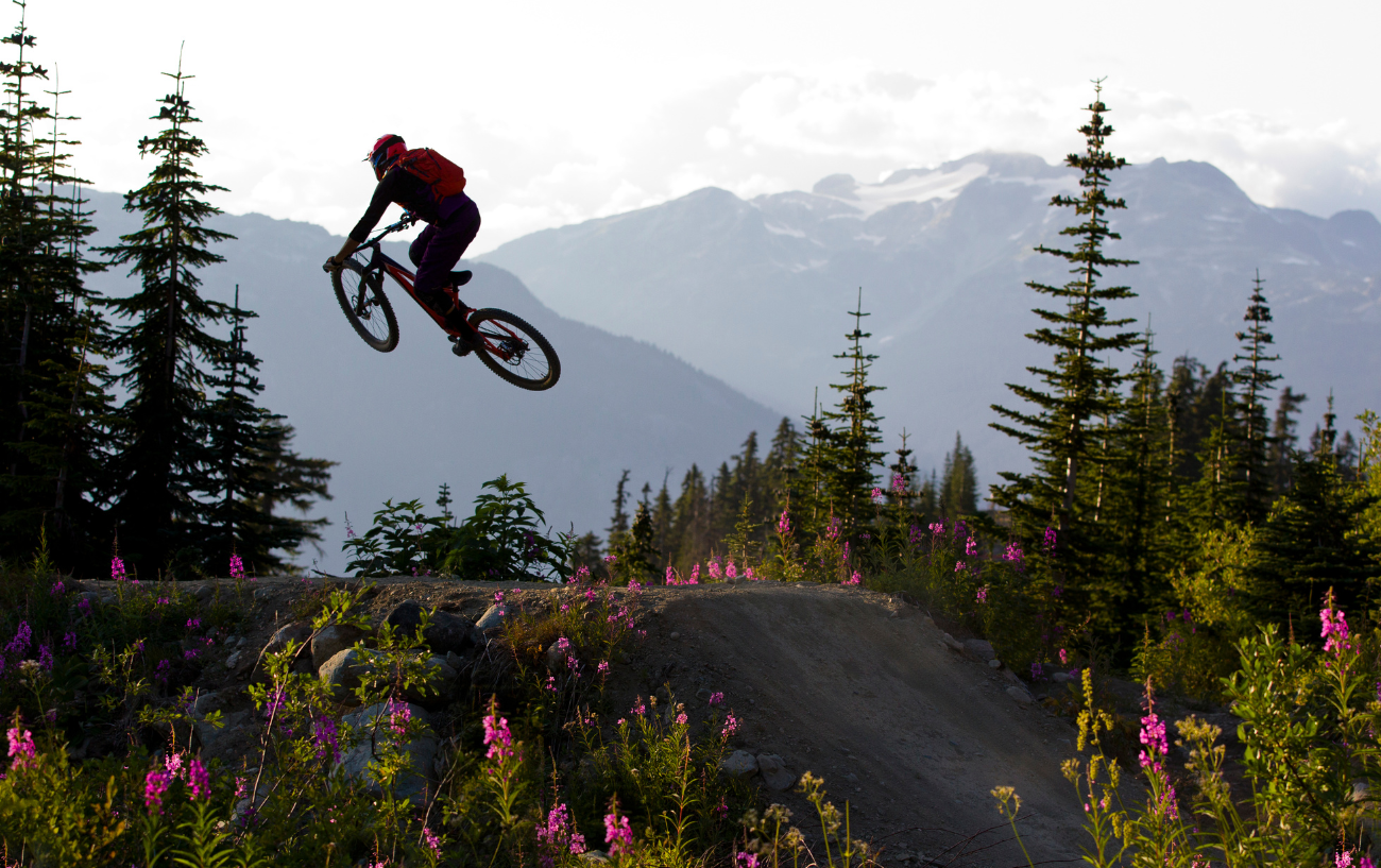 A dirt jump bike leaps over a tabletop feature against a mountainous backdrop.