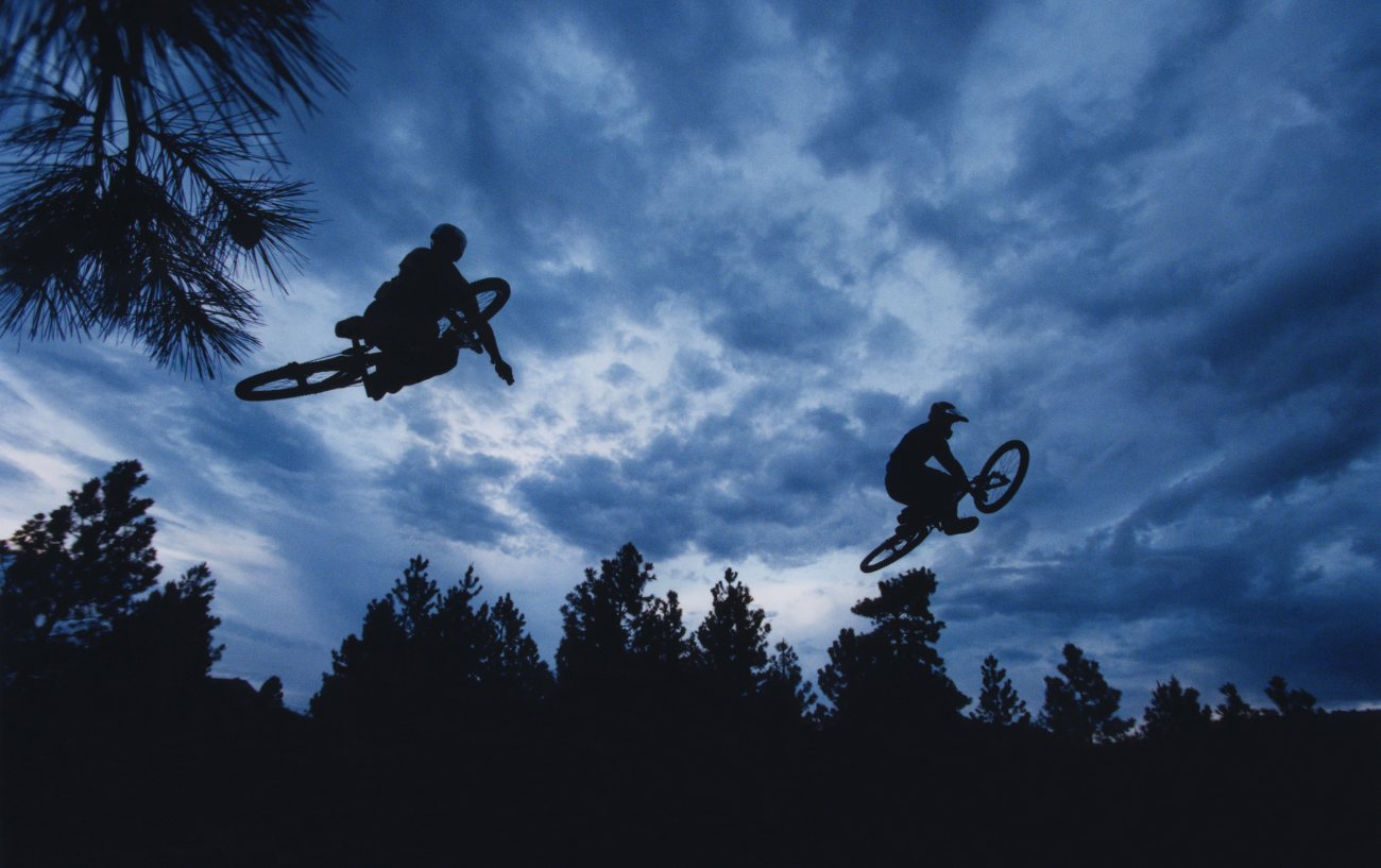 Two silhouetted dirt jump riders perform stunts against a cloudy night sky.