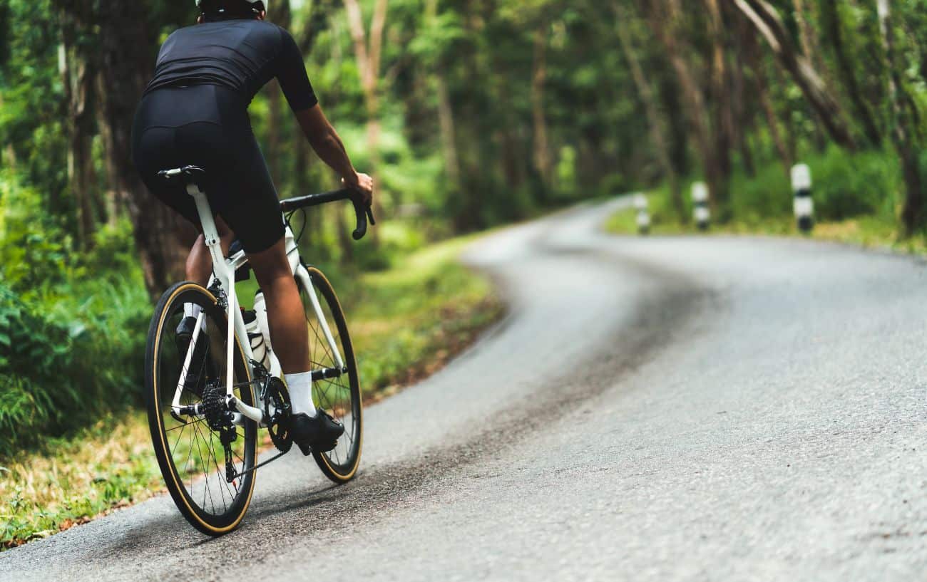 A cyclist on a white road bike climbs a steep tarmac road through a forest.