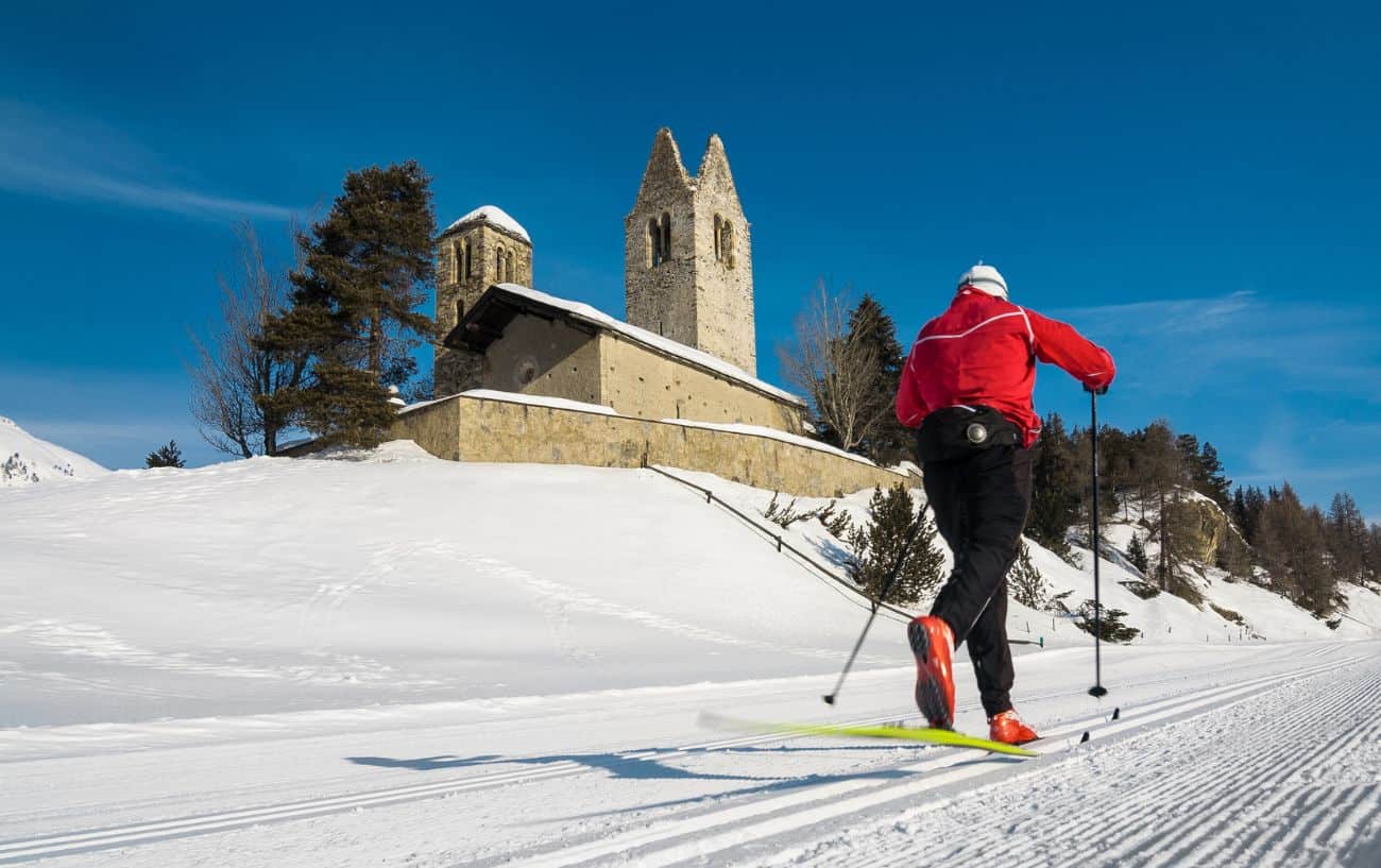 A cross country skier in a red coat passes a snow-covered church.