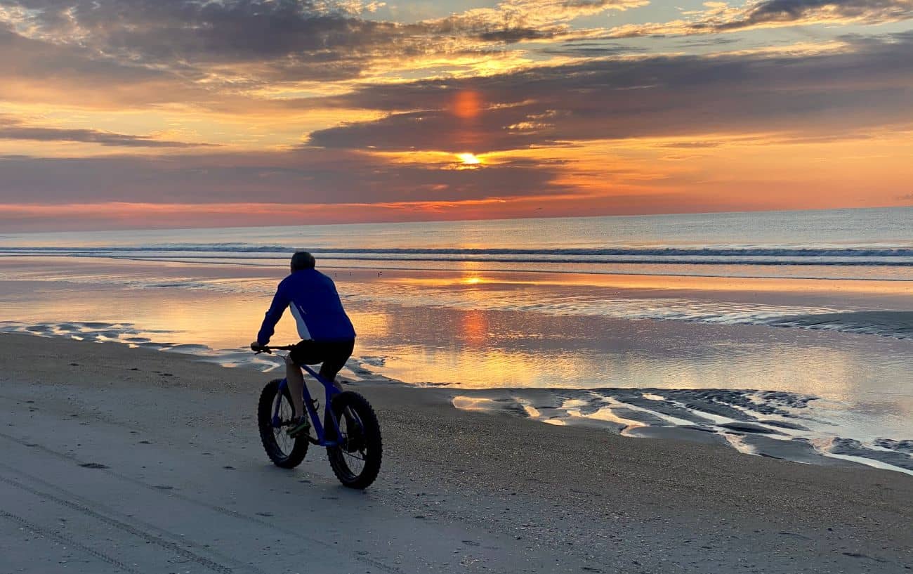 A cyclist rides a fat bike along a sandy shoreline at sunset.