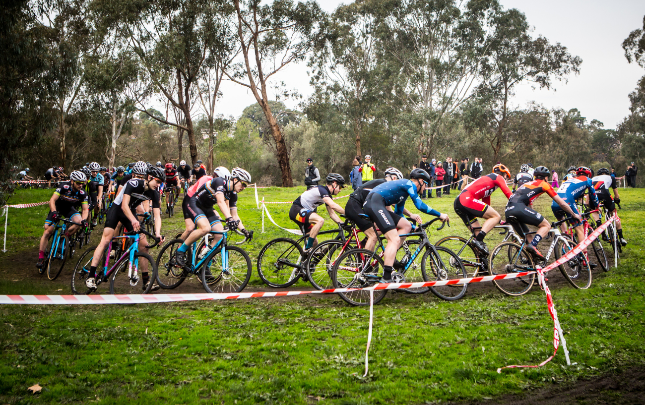 A mass of cyclocross riders tackle a bend on a muddy circuit.