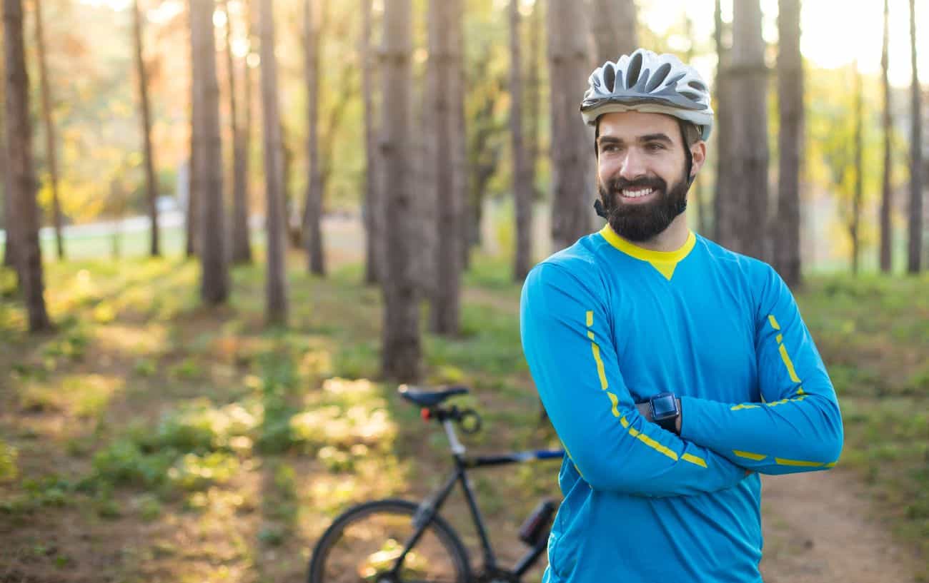 A cyclist in a blue jersey smiles as he stands in a forest clearing.