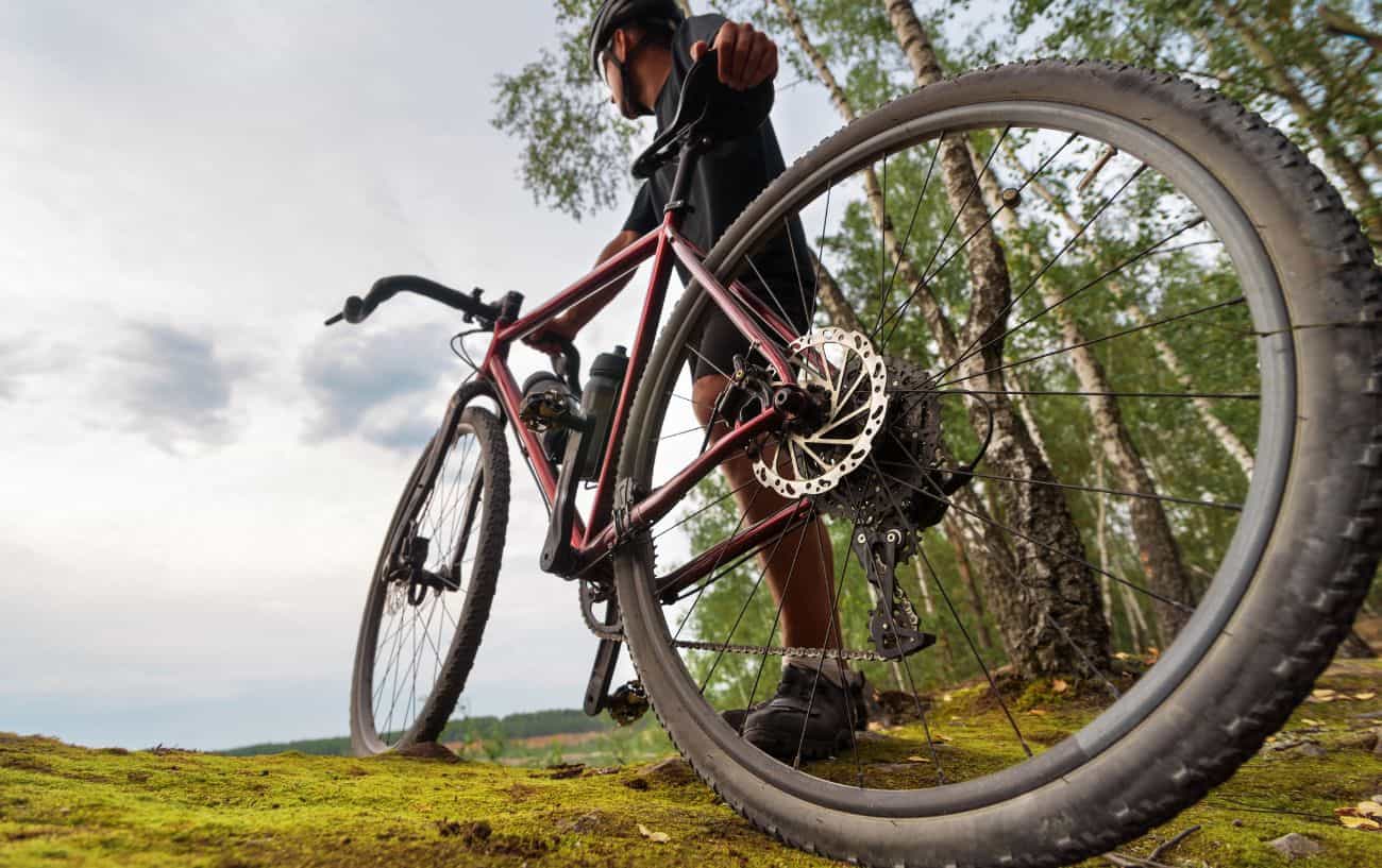A cyclist dressed in black holds a gravel bike on the edge of a forest.