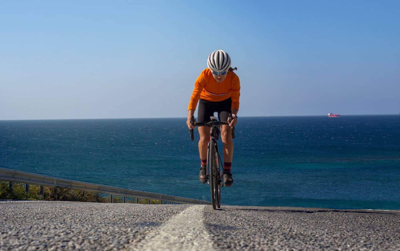A cyclists in an orange jersey climbs a steep coastal road.
