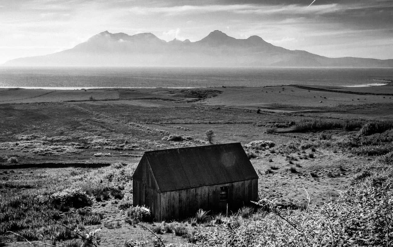 Black-and-white photo of a Hebridean blackhouse, with a mountain range across the water in the distance.