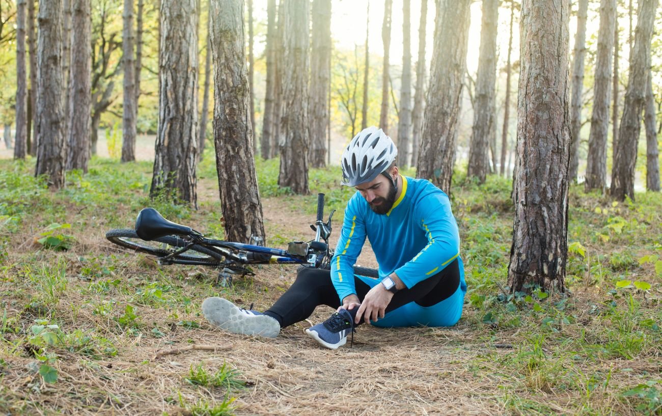 A cyclist in a blue jersey holds his ankle in a forest as he suffers one of the signs of overtraining syndrome.