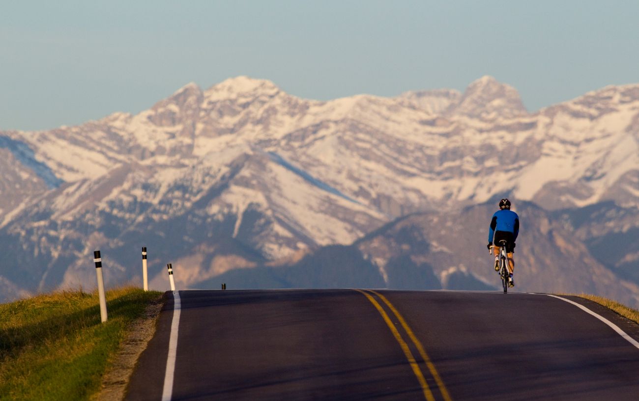 A cyclist reaches the summit of a mountain pass with snow-covered peaks in the background.