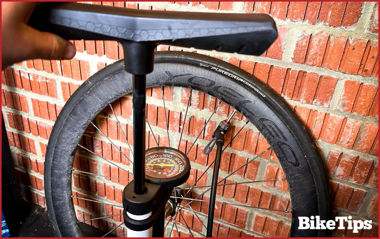 A track pump in front of a deep-section aero bike wheel, resting against a brick wall.