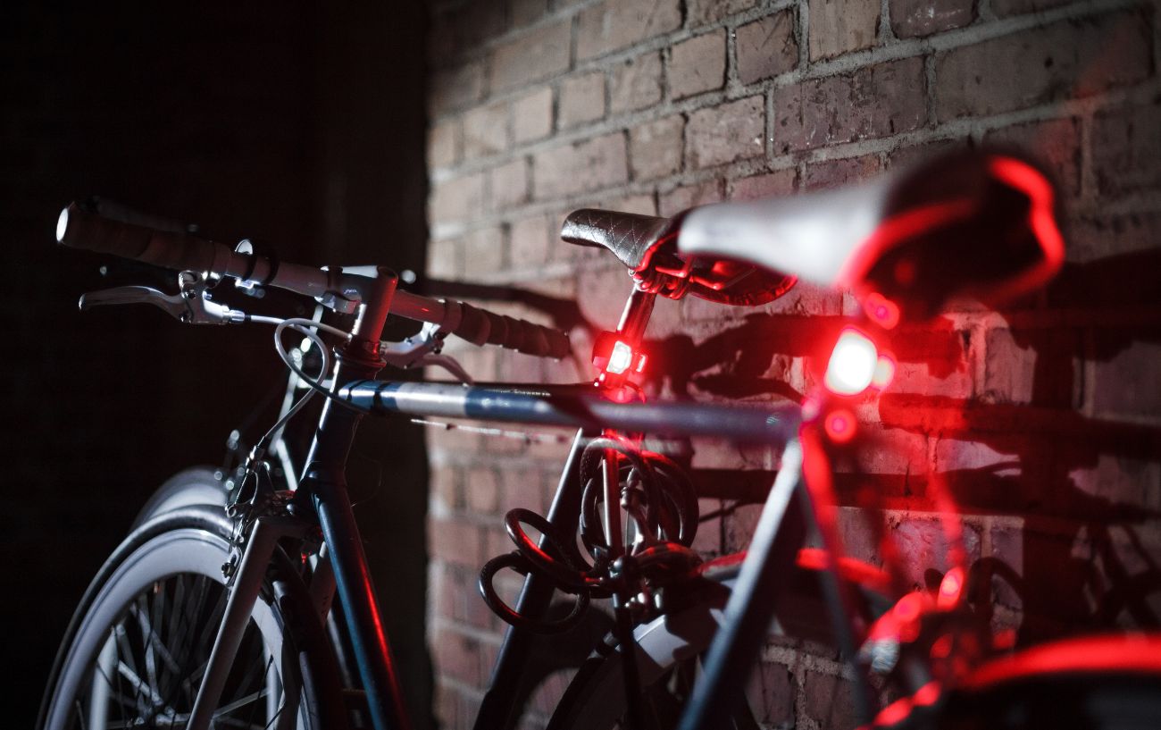 A pair of bikes with red rear lights turned on lean against a brick wall.