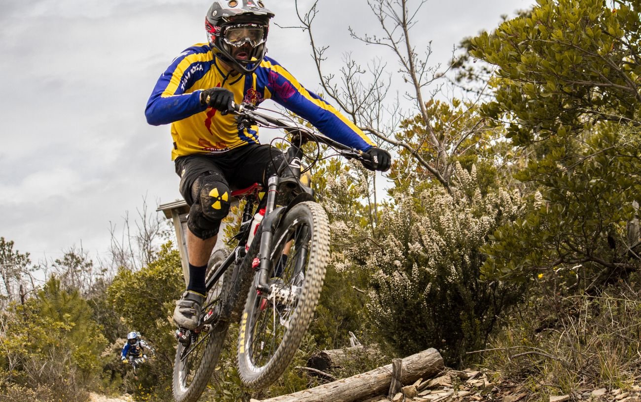 A mountain bike with flat handlebars jumps over a log.