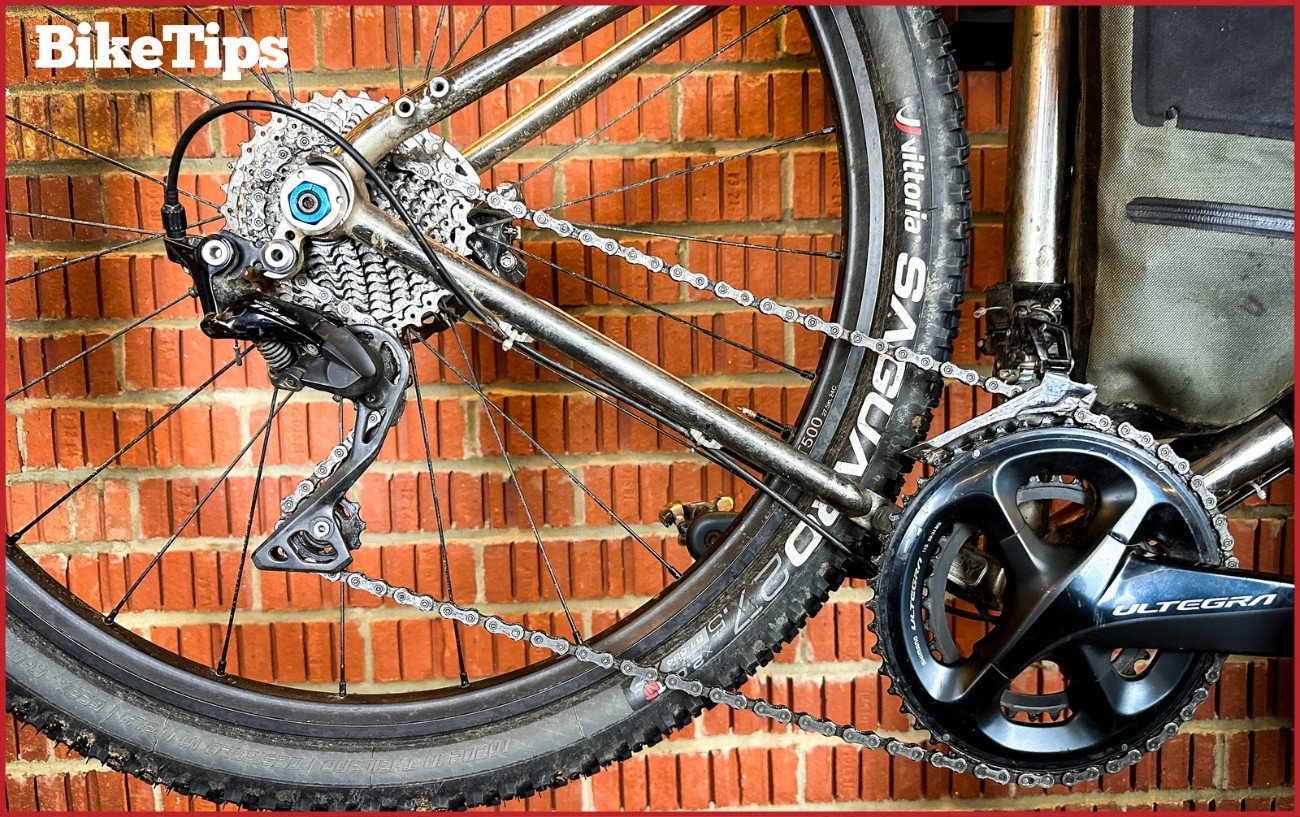 The drivetrain on a silver gravel bike with a raw-brick background.