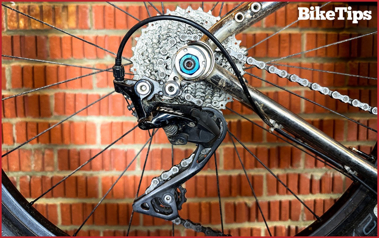 A fully installed rear derailleur of a silver gravel bike on a stand against a raw-brick background.