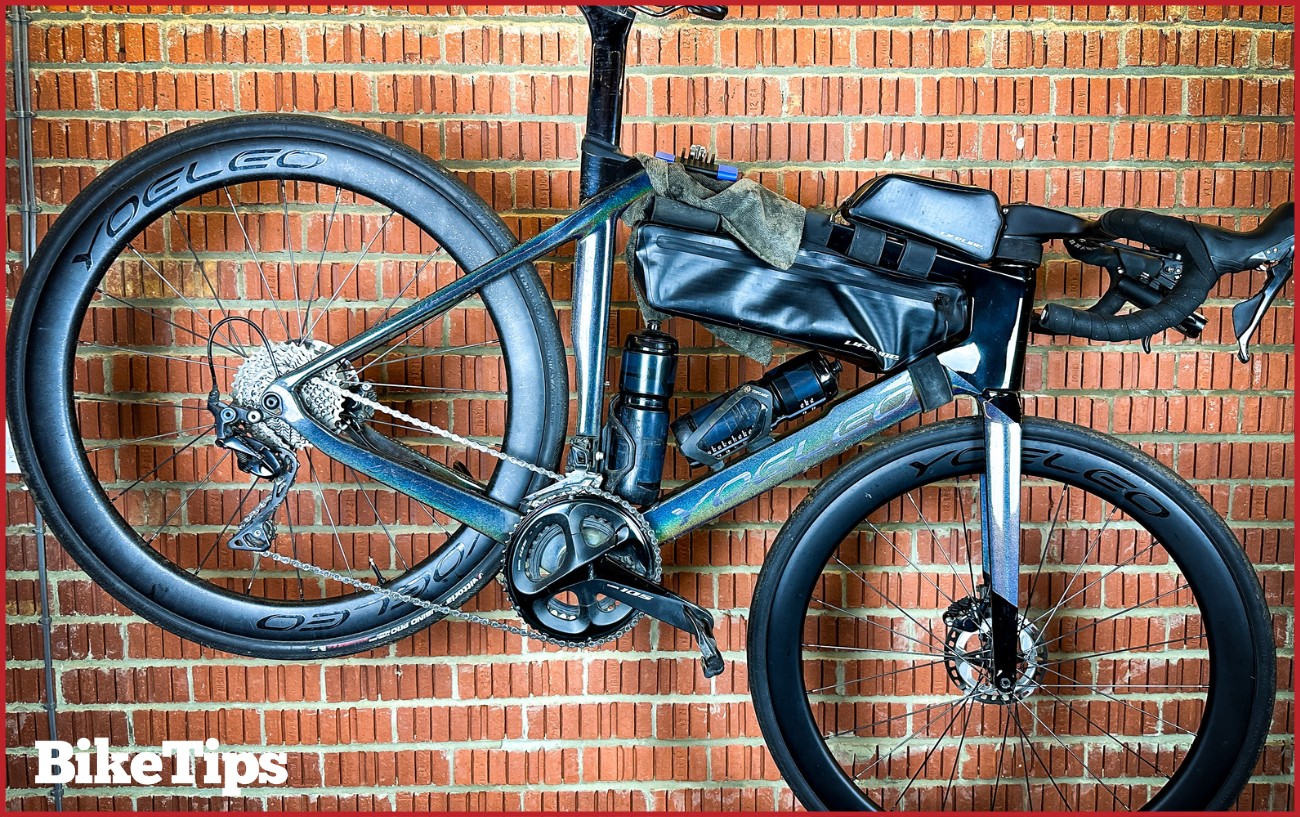 A silver bike sits in a maintenance stand against a brick wall.