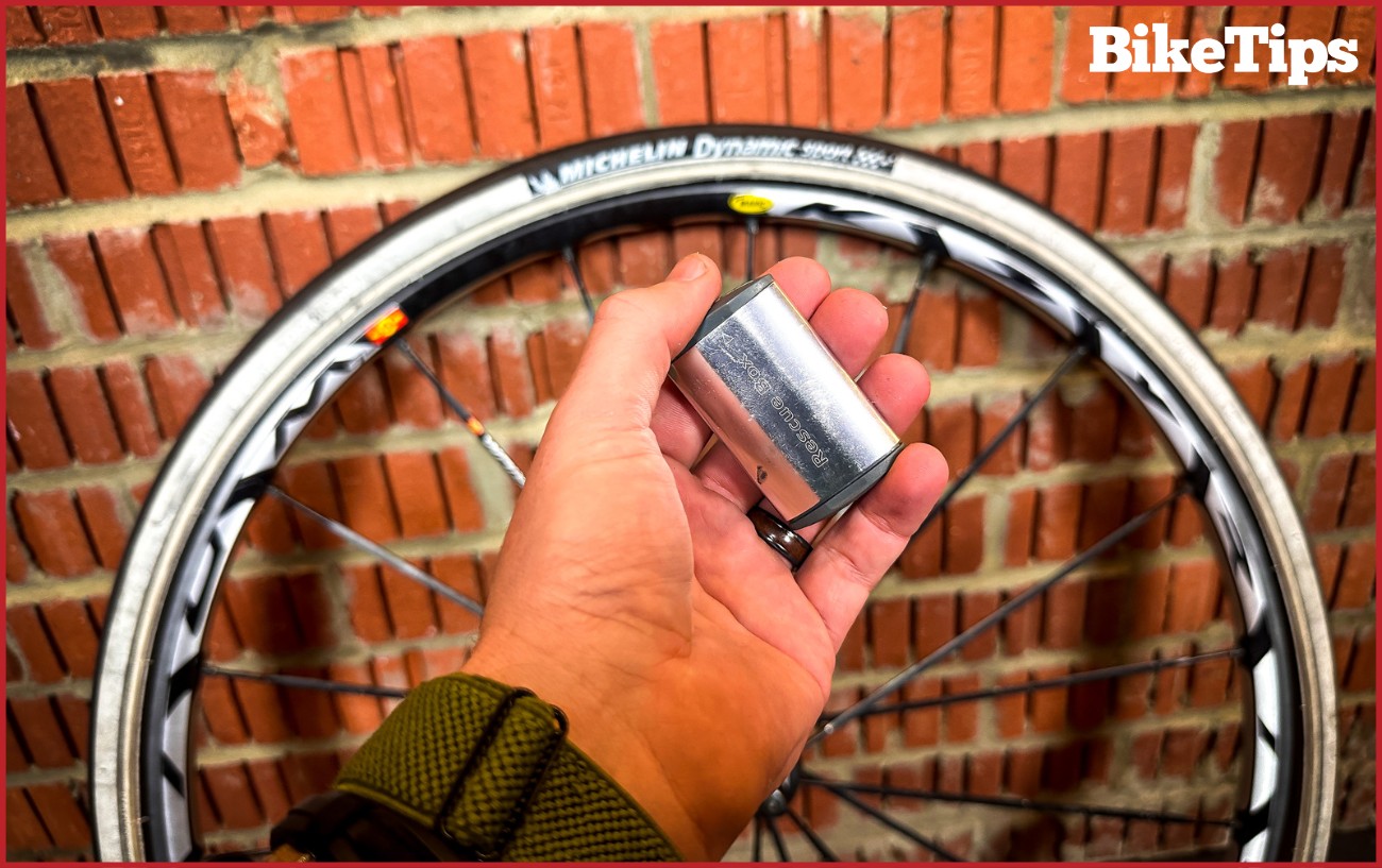 Holding a silver bike patch kit in front of a bike wheel.
